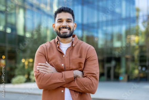 A smiling man stands confidently with arms crossed, posing in front of a modern glass building. The outdoor setting captures a professional and approachable vibe.