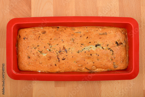 A freshly baked loaf cake with walnuts, raisins, chocolate pieces, and orange zest in a red silicone baking pan on a wooden surface