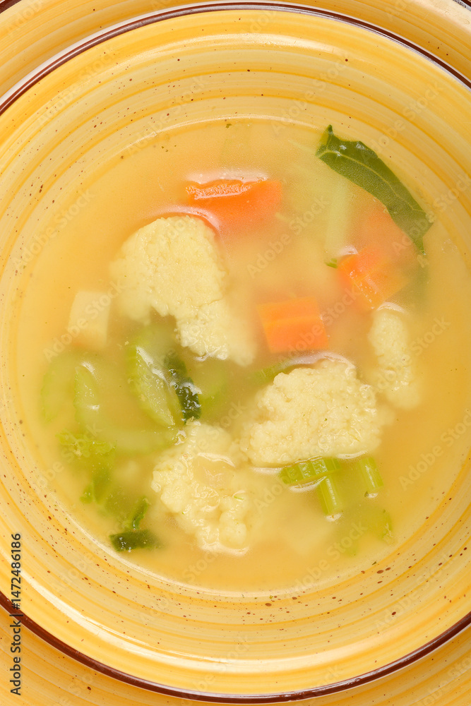 A close-up shows a yellow bowl filled with clear vegetable soup containing semolina dumplings, carrots, and green herbs