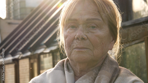 Elderly woman with blonde hair and deep wrinkles on skin is posing with neutral expression on face. Female stands outdoors against of a greenhouse on a summer evening sunset warm light