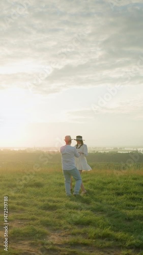 Wallpaper Mural A loving couple spins joyfully on a grassy hill at sunset, beside a calm lake. The husband, in a white shirt and hat, embraces his wife in a flowing white dress, capturing a moment of pure romance Torontodigital.ca