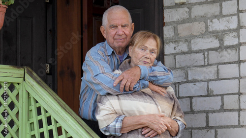 Elderly man in a blue shirt hugging old woman in shawl on their porch. Captures moment of love and affection between couple. Showcasing their bond and happy. Taking care of dear loved ones