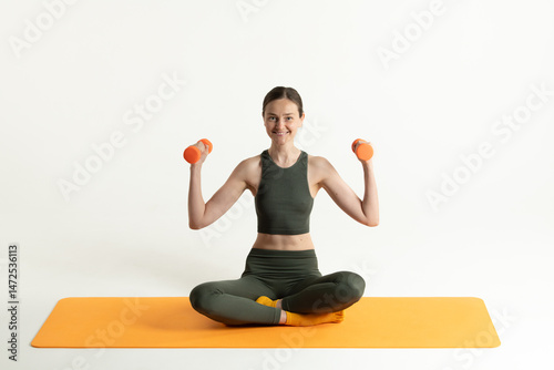 A woman sits cross-legged on an orange mat, lifting small dumbbells. She is in a bright room, showcasing her fitness routine and focus on health