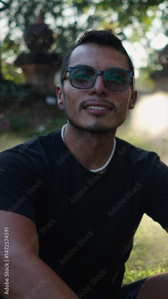 Close-up of the bust of a young Latino man with large glasses smiling at the camera sitting in the garden Close-up of the bust of a young Latino man with large glasses smiling at the camera sitting in the garden