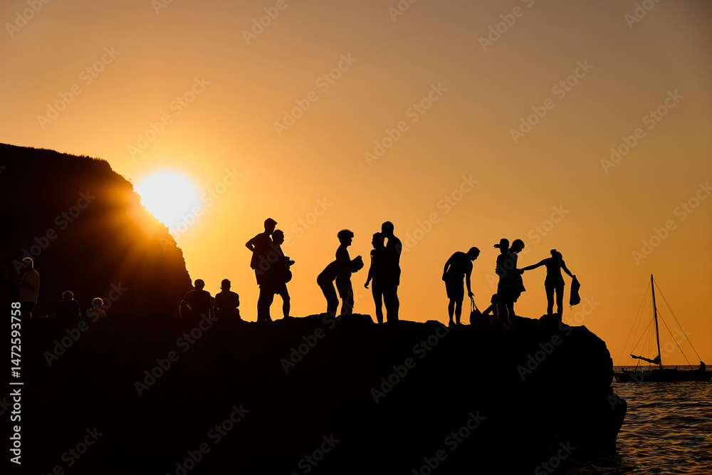 Fototapeta premium Silhouettes of people at sunset on a mountain or cliff, enjoying the view and relaxing in a warm golden light. Summer social moment in nature.
