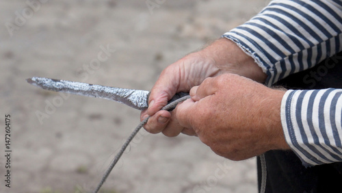 Sailor in striped shirt repairs an old net with knife before fishing. Fisherman holds knife in hand and rope in other, cutting the line. Hobbies and fishing. Close up of elderly mans hands