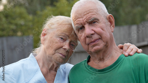 Elderly man and woman stand close together, their faces conveying a sense of solemnity. Female rests hand gently on mans shoulder, while he gazes directly ahead. Love and support of close relatives