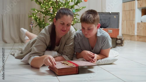 mom and son in casual clothes lying on marble floor opening large vintage book smiling warmly cozy festive indoor setting background featuring christmas tree with lights family moment together