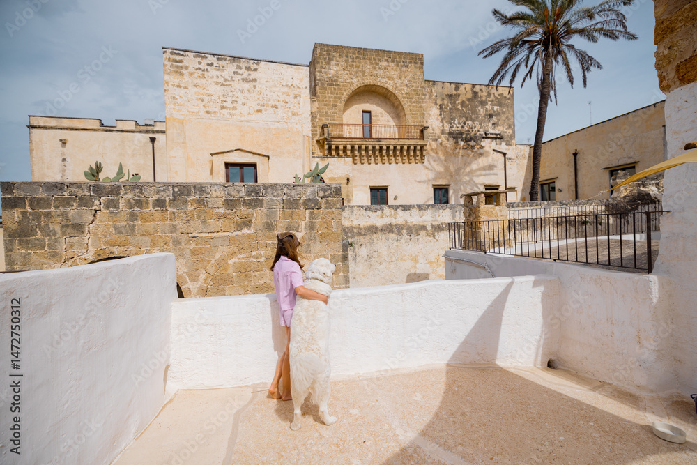 Fototapeta premium A woman and her white dog share a quiet morning moment on a sunlit rooftop, overlooking historic stone buildings and a palm tree in southern Italy