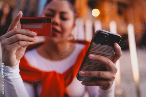 Confident young woman makes a secure online payment with her credit card and smartphone. The vibrant urban setting and warm lights emphasize modern financial freedom and digital shopping convenience