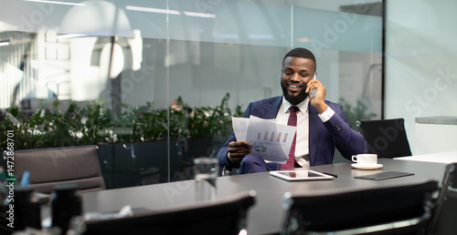 Wealthy black guy in suit entrepreneur having phone conversation with his assistant or business partner, sitting at table with digital tablet on, looking at documents, panorama with copy space