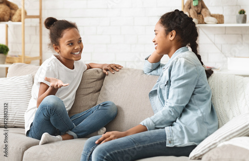 Two lovely african sisters having conversation at home, sitting on sofa at living room, copy space photo
