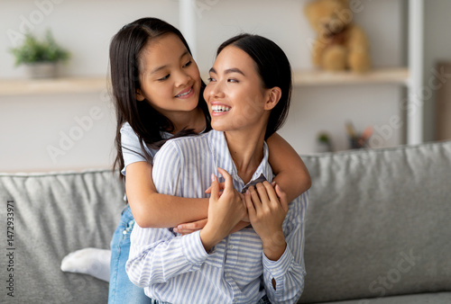Happy asian little girl hugging her beautiful mother and smiling to each other, sitting on sofa in living room, copy space. Adorable family young woman and her daughter embracing