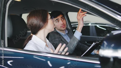 Car dealership conversation between a young man and woman discussing features and options while sitting inside a vehicle during a cloudy day