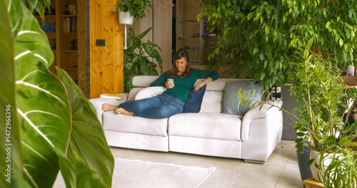 Young lady lounges comfortably on white couch, focused on her smartphone. Filled with lush indoor plants and wooden walls, the cozy space reflects a peaceful, modern lifestyle connected with nature.