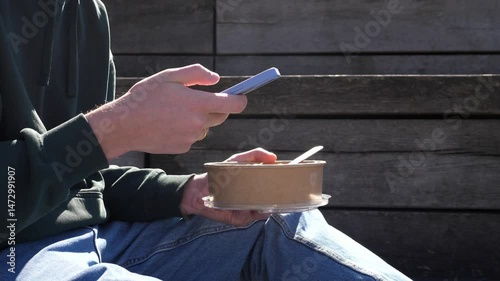 Close-up of a man eating healthy takeaway food and holding a smartphone in his hand, chatting with friends, scrolling through the news feed