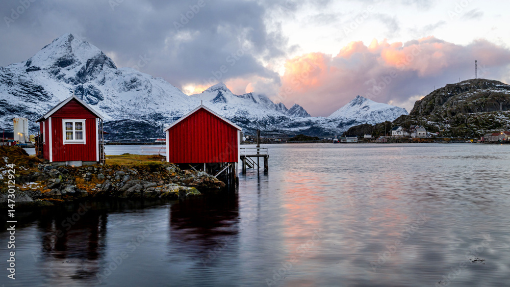 Fototapeta premium A group of traditional red houses, rorbuer, stand in front of crystal clear water and snow-covered mountains, Lofoten, Norway 