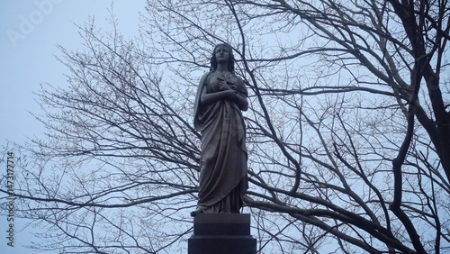 Bronze statue of a woman holding a cross in her hands, surrounded by bare tree branches in a cemetery