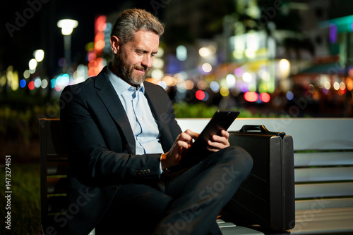 Businessman working on tablet outdoors. Businessman with tablet outdoor. Businessman with a tablet on night street. Businessman using tablet, sit on bench. Night business.