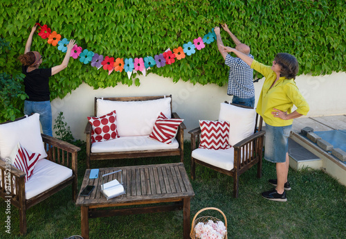 Middle-aged friends hanging colorful floral garland to decorate backyard for outdoor celebration