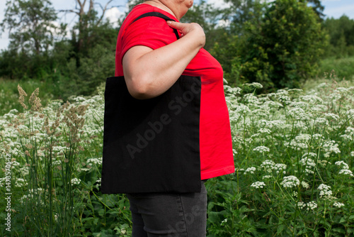 Mature senior woman, white female holding black eco bag canvas fabric for mock up blank template on meadow with flowers background. Ecology, reusable concept. Eco friendly tote shopper