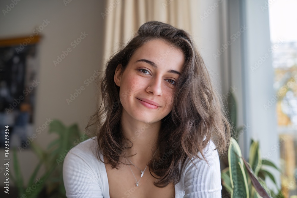 A young woman with long wavy hair smiles warmly while sitting by a window surrounded by greenery. A young woman with long wavy hair smiles warmly while sitting by a window surrounded by greenery.