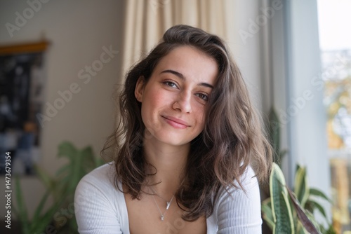 A young woman with long wavy hair smiles warmly while sitting by a window surrounded by greenery. Free Download A young woman with long wavy hair smiles warmly while sitting by a window surrounded by greenery. - GFXLoad