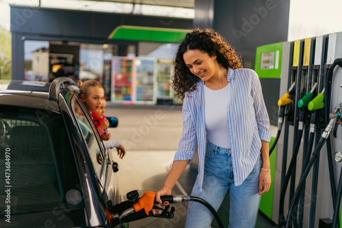 Smiling mother refueling her family car at a gas station, while her two children patiently wait inside the vehicle, enjoying the moment and looking out the window