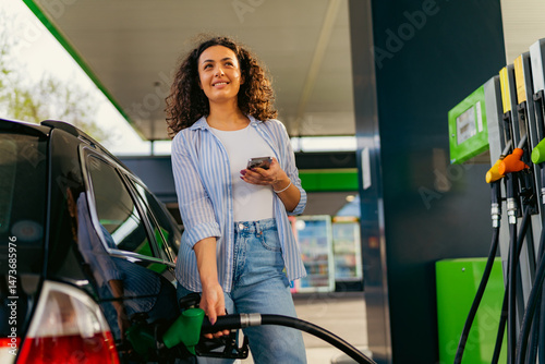 Fotografía Happy young woman refueling her car at a gas station, holding a smartphone in on