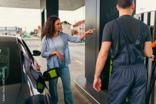 Young woman pointing her finger at something on the gas pump while talking with a gas station attendant, asking for assistance with refueling her car