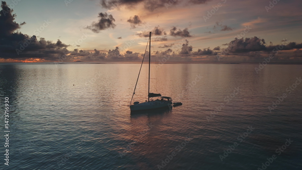 Fototapeta premium Sailboat resting on calm ocean waters of Bora Bora during a vibrant sunset, with colorful clouds reflecting on the water surface, creating a picturesque seascape. Exotic travel vacation getaway
