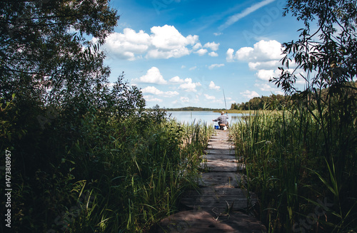 One unrecognizable fishman has fishing peacefully by a calm lake under a blue sky with fluffy clouds. Rear view. Male hobby. Relaxing outdoor activity in a serene natural setting. High quality photo.