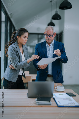 Asian businesswoman and senior businessman collaborating in a modern office, reviewing documents together while analyzing paperwork and discussing strategies at a desk with a laptop
