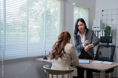 Asian business manager pointing at her watch while scolding employee for being late at the office, concept of discipline and punctuality in business