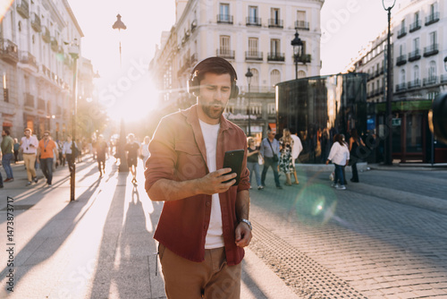Fotografija young man listens to music with wireless headphones while reading something on his smartphone in crowded city square