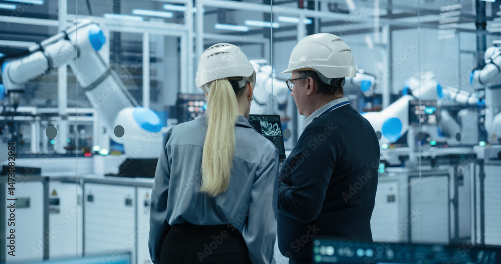 Obraz premium Electronics Factory: Female Project Manager and Engineer in Hard Hats, Talking, Using Laptop. Monitoring Advanced Equipment Production. Robotic Arms Assembly Line Manufacturing Circuit Boards