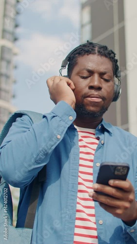 Smiling African American male enjoying music through headphones while checking phone outdoors. Holding one side of headset and reacting to lyrics. Walking in city and browsing playlist updates.