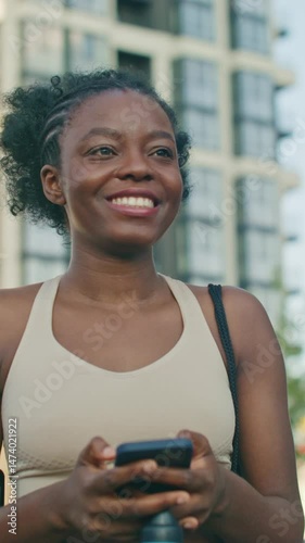 Cute African American female in light sports bra walking outdoors with bottle and smiling at smartphone screen. Texting friend or reacting to message. Enjoying fresh air after finishing fitness.
