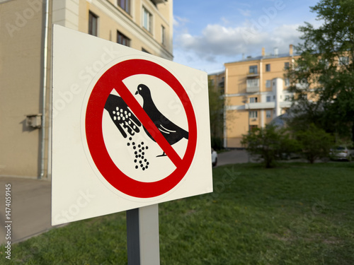 Warning sign in residential courtyard prohibiting feeding pigeons. Simple urban design with clear pictogram against blurred background of apartment buildings and green lawn