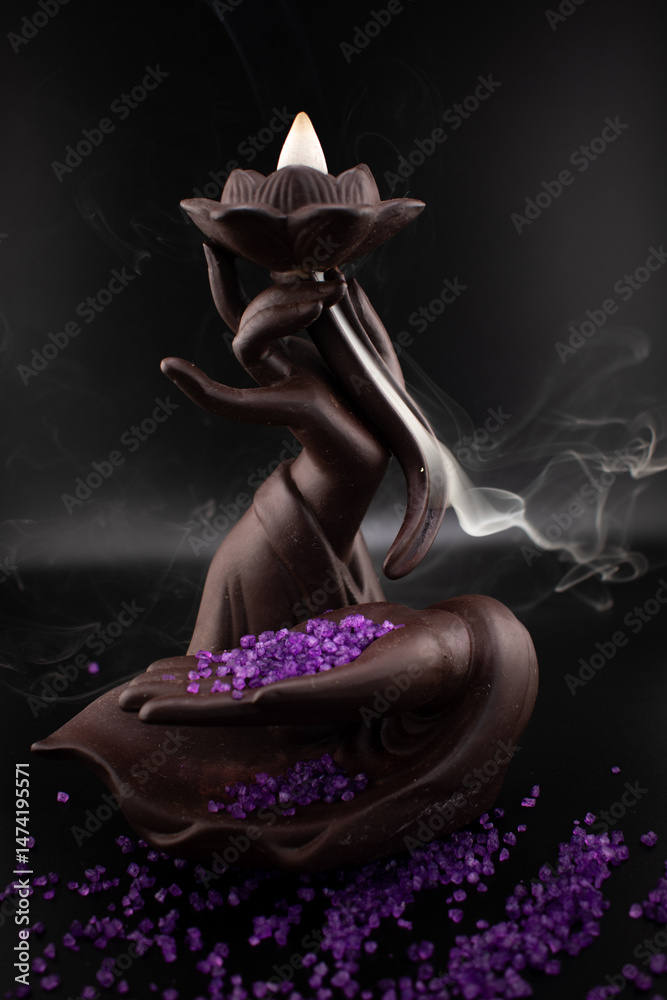 Obraz premium Ethereal scene of a backflow incense burner shaped as a hand holding a lotus with fragrant smoke cascading downwards amidst scattered purple sea salt Evokes mystery, tranquility and spiritual ambiance