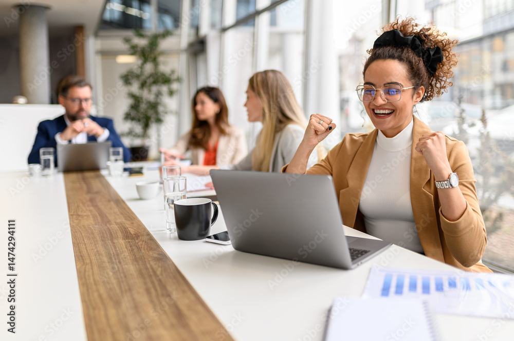 Obraz premium Businesswoman shaking fists and celebrating success during online meeting in office with colleagues in the background