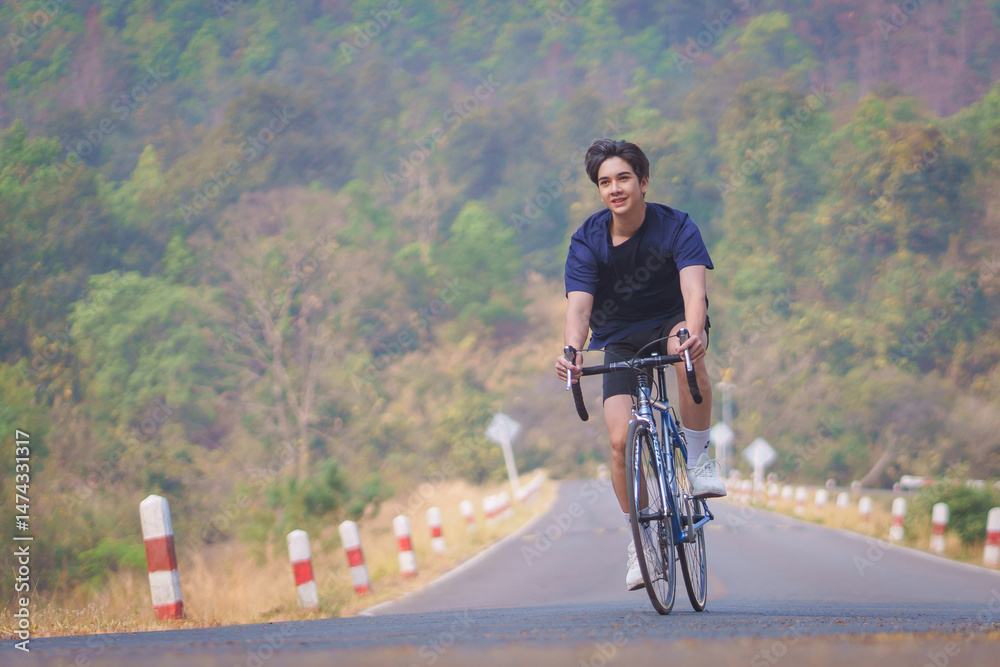 Fototapeta premium Handsome young man in a blue shirt and black sports shorts is happily riding a bronze and blue bicycle in the morning for exercise in the park during cold holiday weather. Healthy exercise concept.