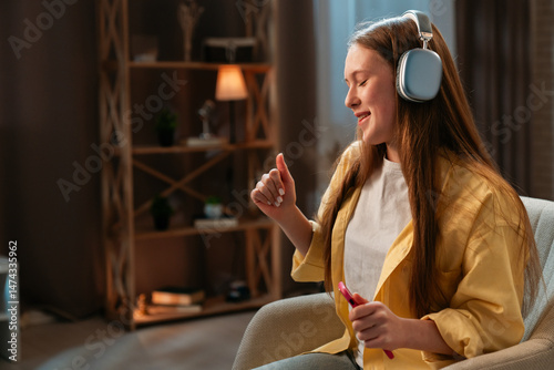 Young teenager girl spending evening time at home, girl sits in chair in the living room and listening music in headphones, dancing positive expression.