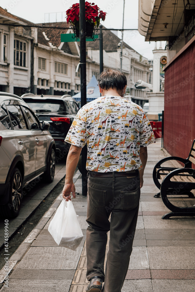 Obraz premium Man walking on city sidewalk carrying plastic bag, rear view