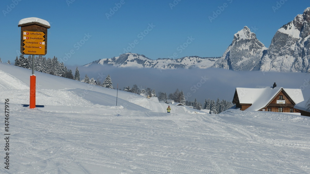 Fototapeta premium Scenic winter landscape: snow-covered mountains, ski slope, cozy cabin, and a trail sign.