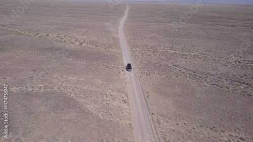A black SUV is driving fast on a dusty road in the steppe. Clouds of dust fly out from under the wheels and from the roof of the car. The car easily overcomes a dirt road. Blue sky and white clouds.