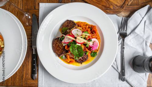 from an overhead angle, a composed dish is presented in a wide rimmed, plain plate. it features dark, round portions nestled against a vibrant sauce, topped with sliced radishes and green leaves