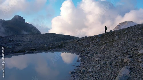 Traveler with a backpack walks along a rocky mountainside path near a small pond, surrounded by clouds reflecting in the water. Drone tracking at evening. Extreme sport adventure and healthy lifestyle