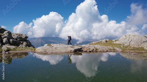 Traveler with a backpack walks along a rocky mountainside path near a small pond, surrounded by clouds reflecting in the water. Drone tracking at evening. Extreme sport adventure and healthy lifestyle