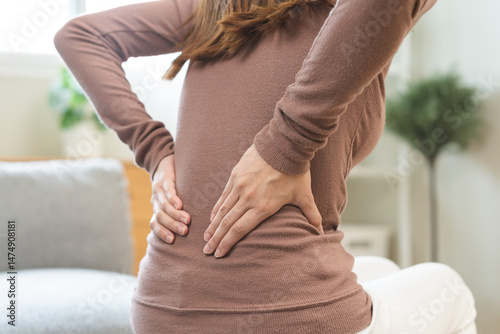 Foto Pain body muscles stiff problem, suffer asian young woman painful back, symptom injury ache from long study work, hand holding massaging or rubbing lower back hurt, sore sitting on sofa at home